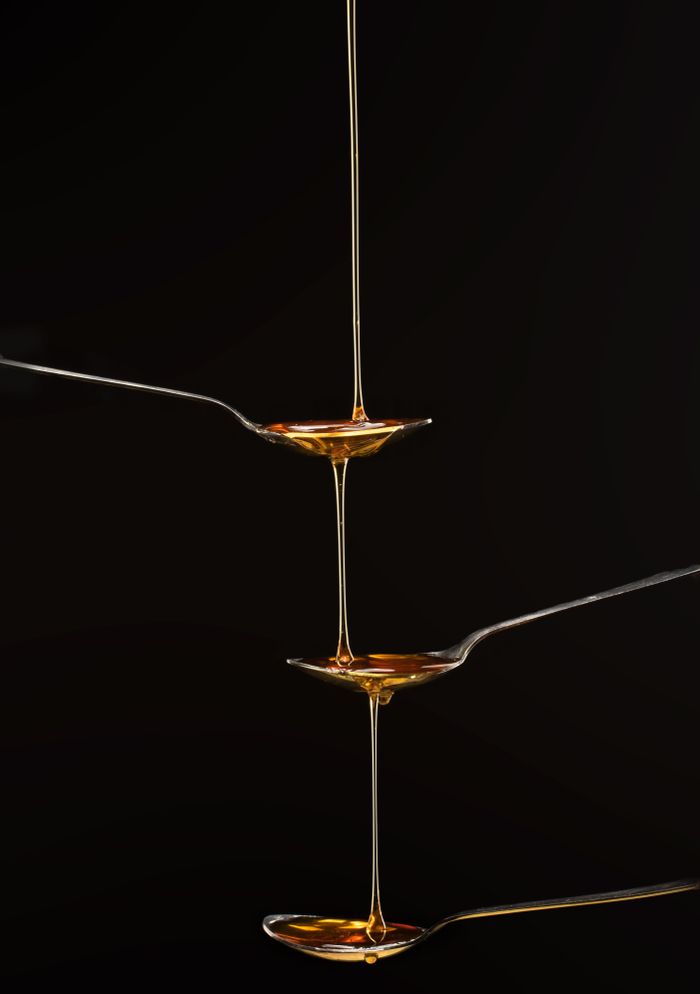 Artistic close-up of golden honey streaming from three aligned spoons on a black background.
